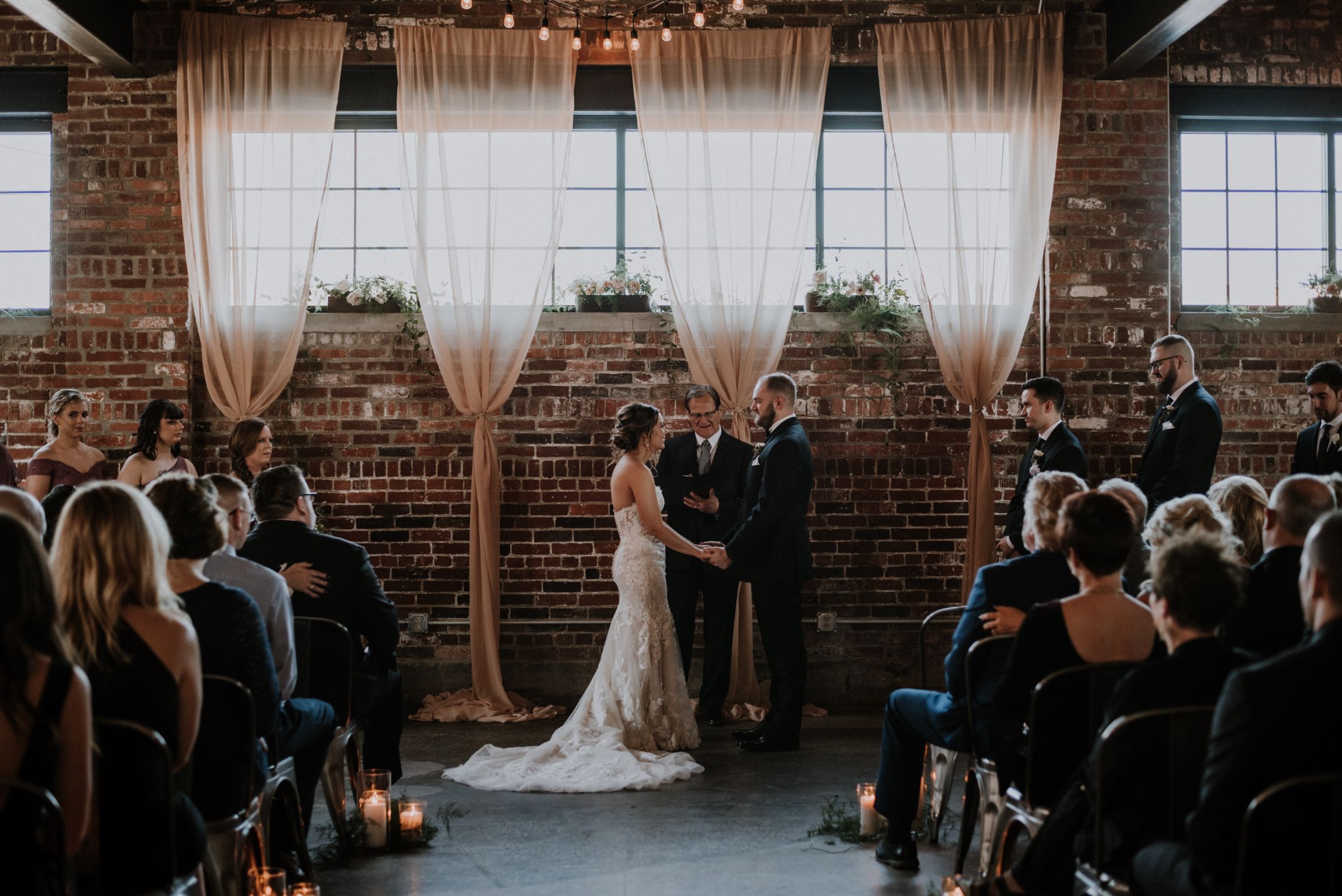 Wedding ceremony with bride and groom holding hands in front of windows and drapery