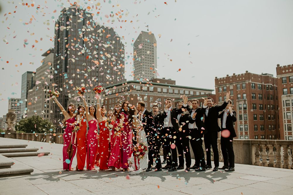 A wedding party throws pink and blue confetti into the air on their rooftop patio.